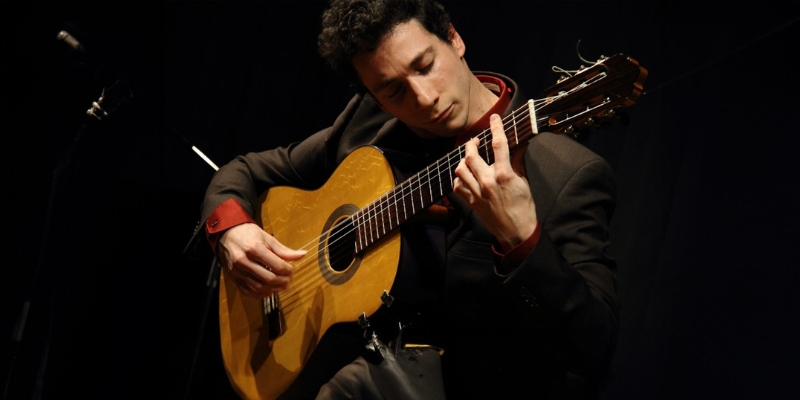 Grisha Goryachev plays the guitar in front of a black background, wearing a dark suit and red dress shirt underneath.