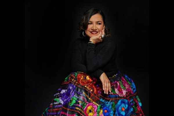 Martha Gonzalez sits and poses in front of a black background, wearing a black turtleneck and a colorful patterned skirt.