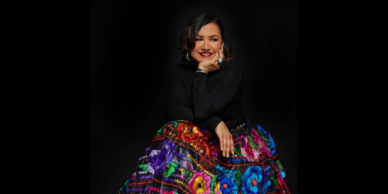 Martha Gonzalez sits and poses in front of a black background, wearing a black turtleneck and a colorful patterned skirt.