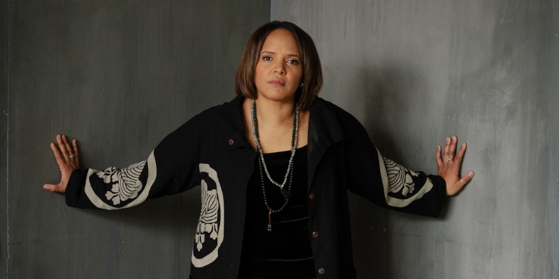 Terri Lyne Carrington standing with her hands placed against two walls, wearing a black jacket with patterned sleeves and a beaded necklace.