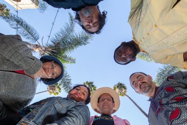 A group image of Quetzal, with all 6 members looking downwards at the camera. A clear blue sky in the background with palm trees.