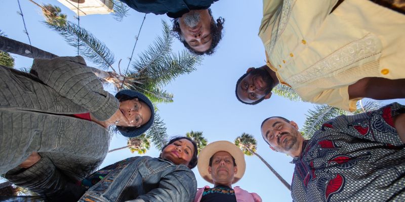 A group image of Quetzal, with all 6 members looking downwards at the camera. A clear blue sky in the background with palm trees.