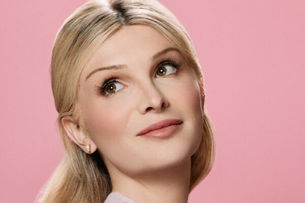 Headshot of Dylan Mulvaney (with fair skin and long blonde hair) posing in front of a pink background
