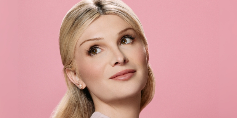 Headshot of Dylan Mulvaney (with fair skin and long blonde hair) posing in front of a pink background