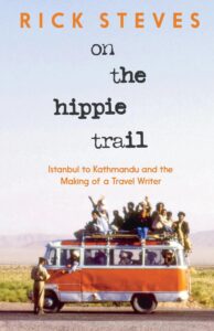 Book cover of "On the Hippie Trail" by Rick Steves, featuring a vintage orange-and-white van packed with travelers on the roof, parked in a desert landscape under a clear sky. A police officer stands on the road, talking to the driver from the window. Subtitle reads "Istanbul to Kathmandu and the Making of a Travel Writer."