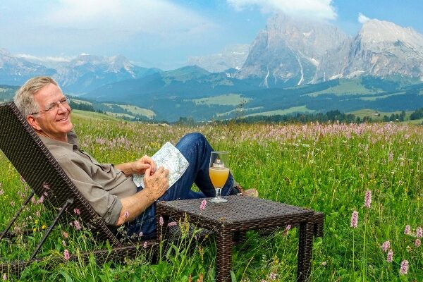 Rick Steves (with short blonde hair & eyeglasses) sits on a wicker chair and holds a paper map, smiling. A matching table is next to him, with a drink on top. The background shows a meadow with cloudy blue sky and mountain landscape in the distance.
