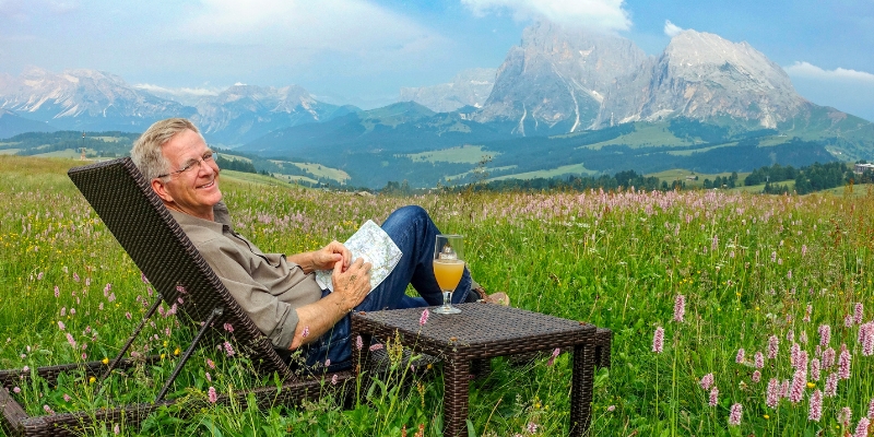 Rick Steves (with short blonde hair & eyeglasses) sits on a wicker chair and holds a paper map, smiling. A matching table is next to him, with a drink on top. The background shows a meadow with cloudy blue sky and mountain landscape in the distance.