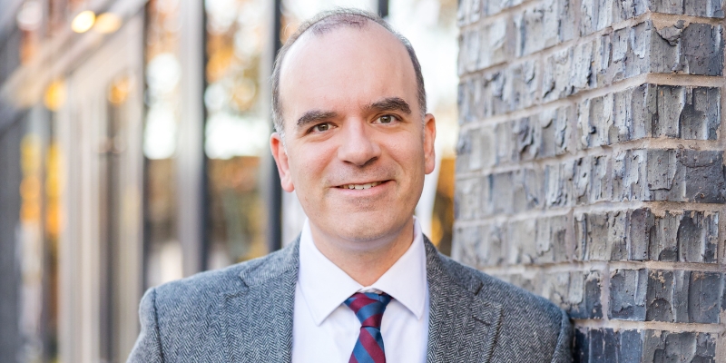 Headshot of Yoni Appelbaum (with fair skin, gray suit, and navy/red tie) standing in front of a brick building with glass windows.