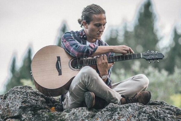 Calum Graham sits on a rock outdoors and tunes his acoustic guitar.