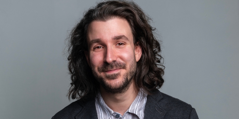Headshot of Adam Met (with fair skin, chin-length brown hair, and beard) posing in front of a grey backdrop.