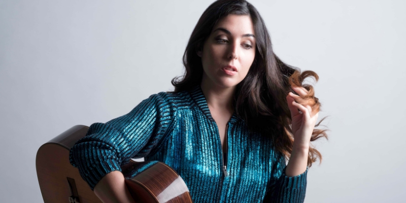 Andrea Gonzalez Caballero poses in front of a white background, holding a guitar and wearing a blue sequin jacket.