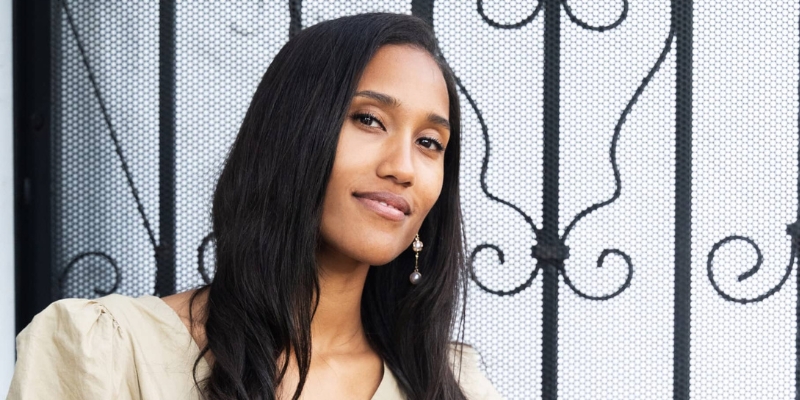 Anna Malaika Tubbs (with brown skin, long black hair) wears a tan blouse with pearl earrings as she poses in front of an iron gate.