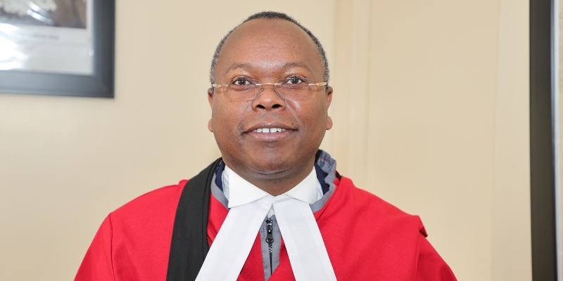 Headshot of Judge Joel Ngugi (with brown skin, grey buzzed curly hair) wearing wireless rim glasses and a red jacket.