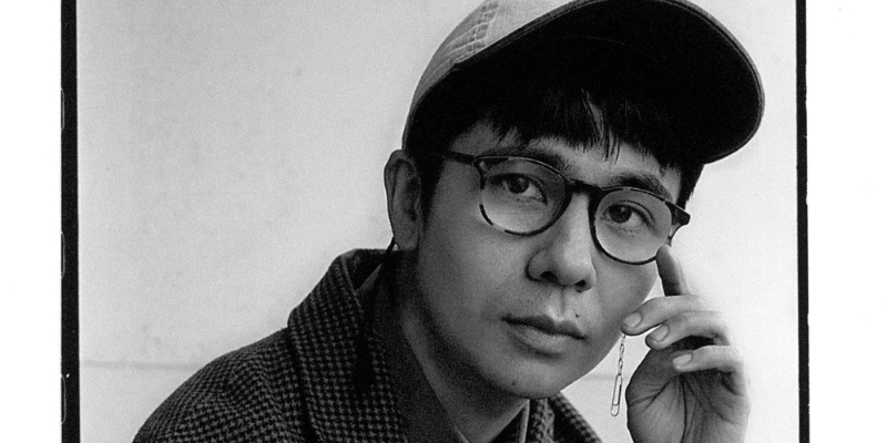 Black and white headshot of Ocean Vuong (with light skin and short black hair, wearing a baseball cap, glasses, and a paperclip earring).