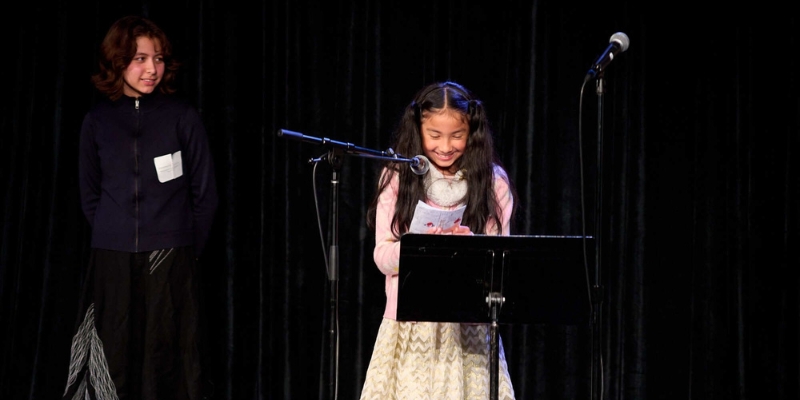 A student with long dark hair and a pink cardigan reads a piece of writing off of a music stand with a microphone in front. Another student watches in the background, wearing a name tag.