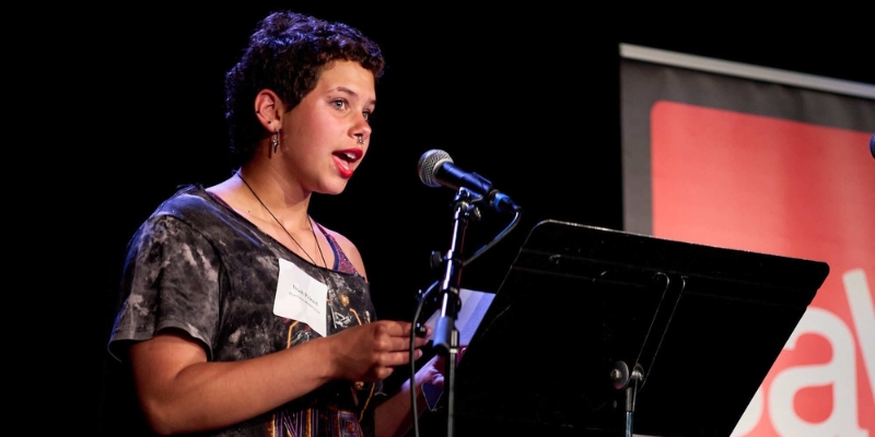 A student with short wavy hair, septum piercing, and red lipstick reads a piece of writing off of a music stand with a microphone in front.