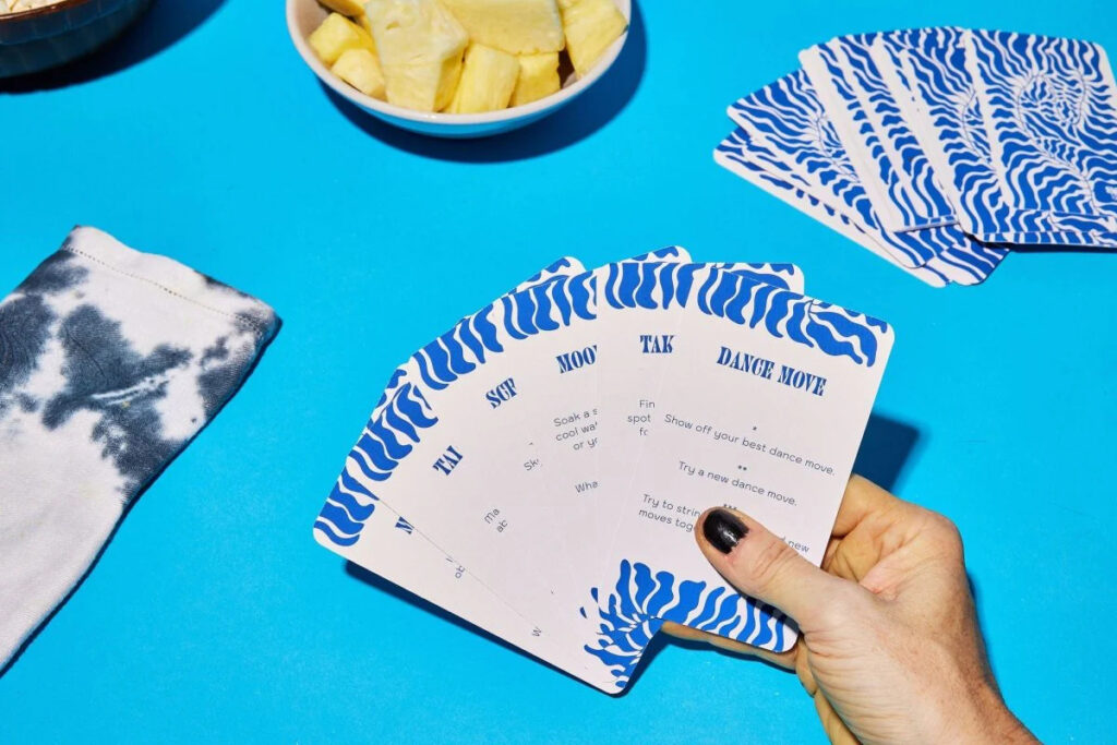 A hand holds a desk of SetSet cards on a bright blue table with other miscellaneous items (a bowl of fruit, a shirt sleeve) nearby