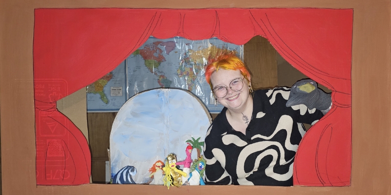 Mikki Ulaszewski (with short orange hair and glasses) smiles and poses behind a painted cardboard stage curtains with various finger puppets.