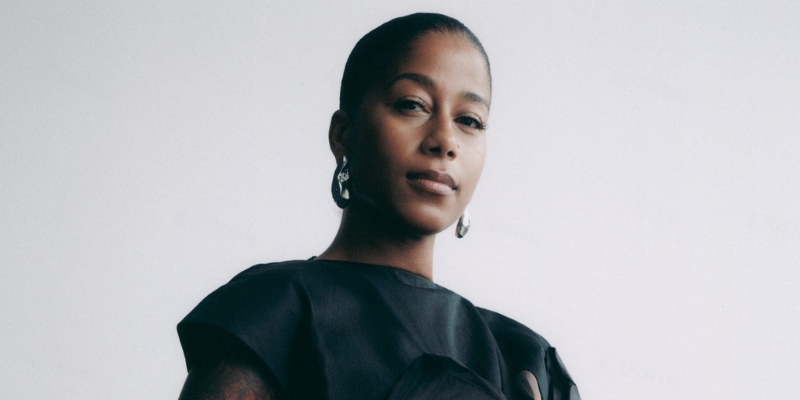 Headshot of aja monet (with brown skin, low black bun, wearing a black blouse and large silver earrings)