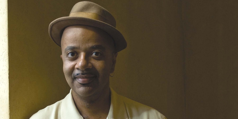 Headshot of James McBride (with brown skin, tan fedora, and beige polo shirt)