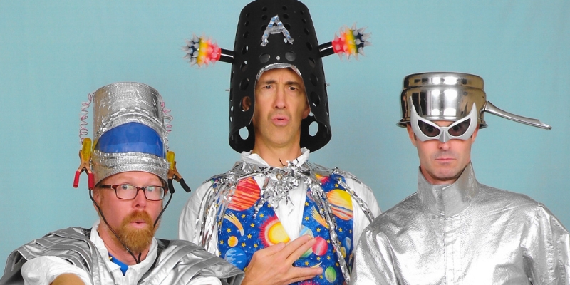 The three members of The Brian Waite Band pose in front of a blue background wearing silver alien/space themed costumes.
