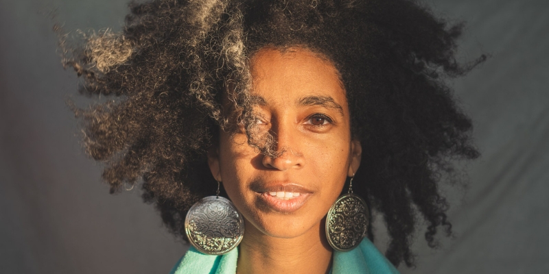 Headshot of Alexis Pauline Gumbs (with brown skin and textured black hair, wearing a blue scarf and large medal earrings).