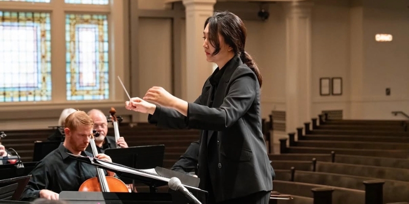 A person with a black ponytail and black suit conducts an orchestra with empty pew seating in the background.