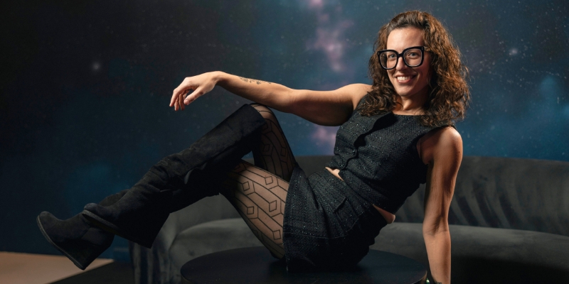 Cate Osborn (wearing a black dress, boots, and large glasses) poses on a black coffee table with one knee propped up. The background is a photo of outer space.