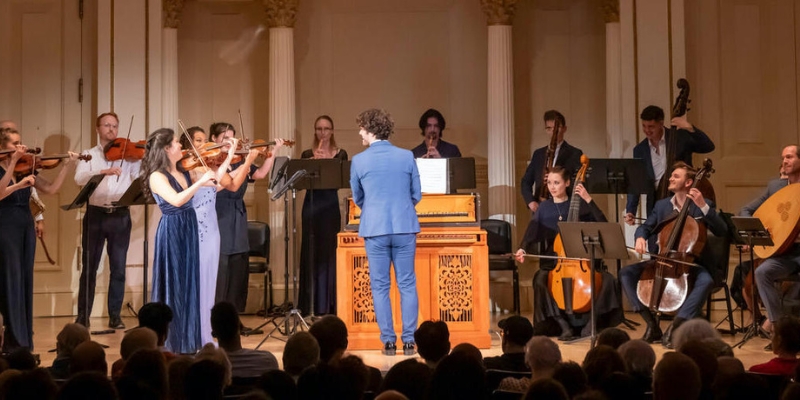 A conductor in a blue suit stands behind a wooden organ and conducts a string ensemble.