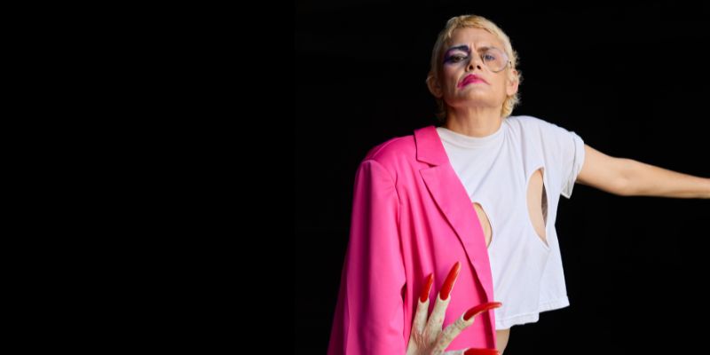 Dynasty Handbag poses with one arm extended and a pink blazer hanging off her shoulder. She has short blonde hair and wears a monocle.