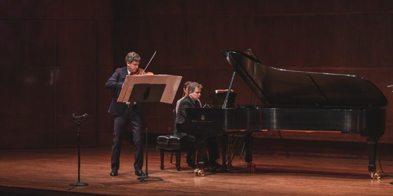 James Ehnes (playing the violin) performs on stage with Andrew Armstrong (playing the piano). Both performers are wearing black suits.