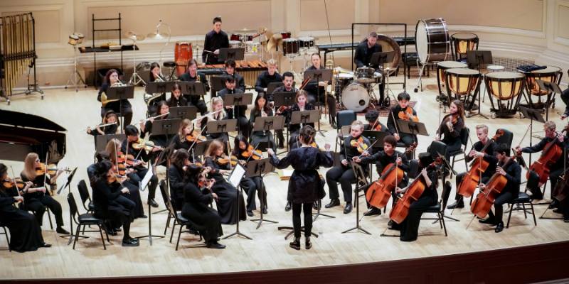 A youth orchestra and conductor perform on a brightly lit stage, all dressed in black.