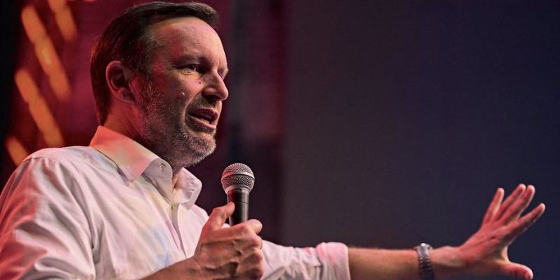 Senator Chris Murphy (wearing a white button-down) gestures with one hand while speaking into the microphone in the other hand.