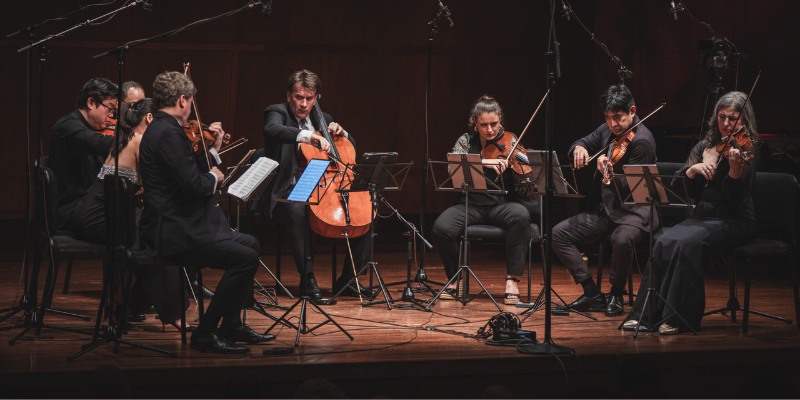 A co-ed string ensemble of 8 performs on a dimly lit stage, dressed in black.