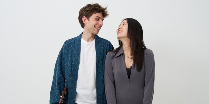 Antoine Boyer and Yeore Kim stand side by side, smiling and looking at each other. Antoine holds the top of an acoustic guitar in one of his hands.