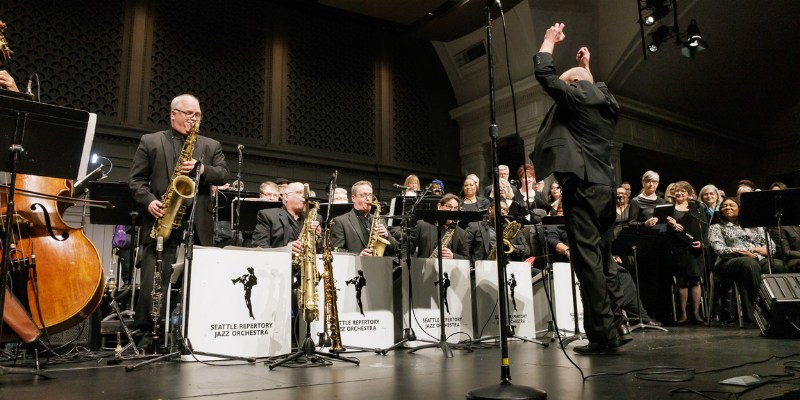 A jazz orchestra performing on stage with musicians playing various instruments, including saxophones and a double bass. The conductor is leading the performance.