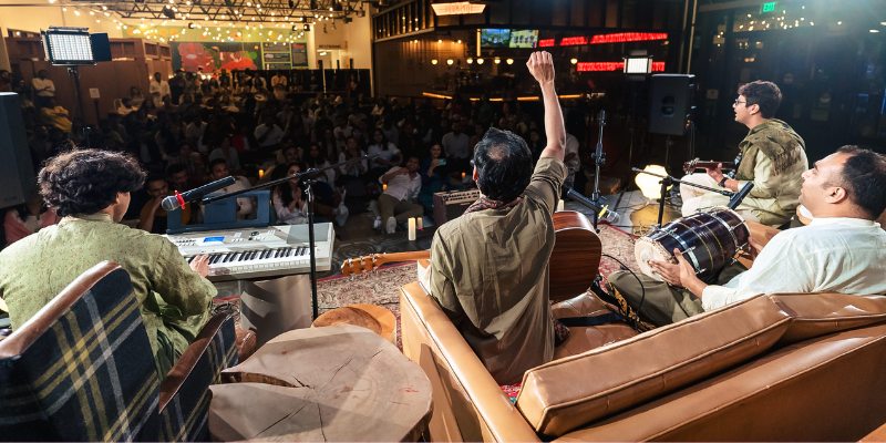 A group of four musicians performs on stage in a lively indoor venue with an engaged audience seated in the background.