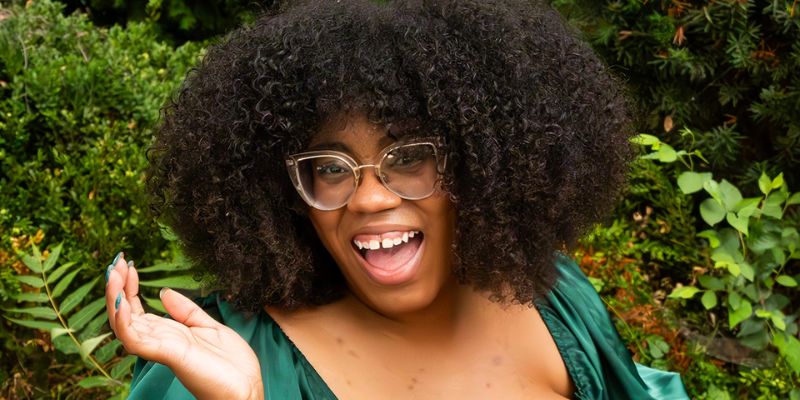 A person with dark, afro hair and glasses smiles with their mouth open, wearing a green satin top, with green foliage in the background.