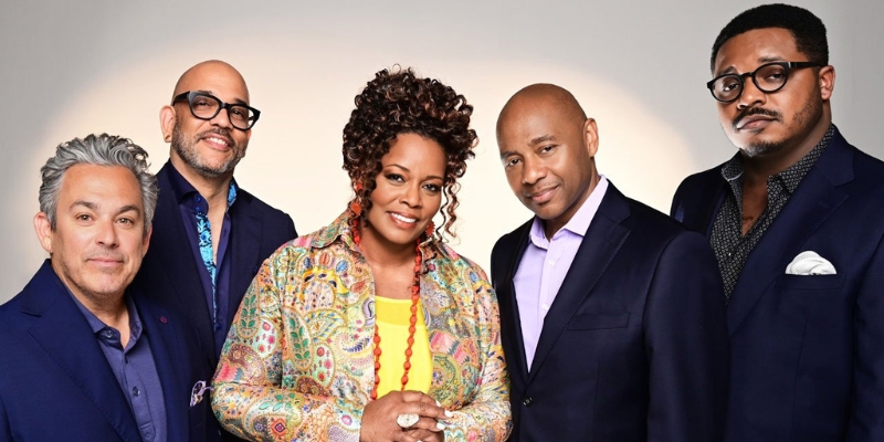 Five adults pose together in a studio portrait, with a smiling woman in a colorful jacket standing at the center.