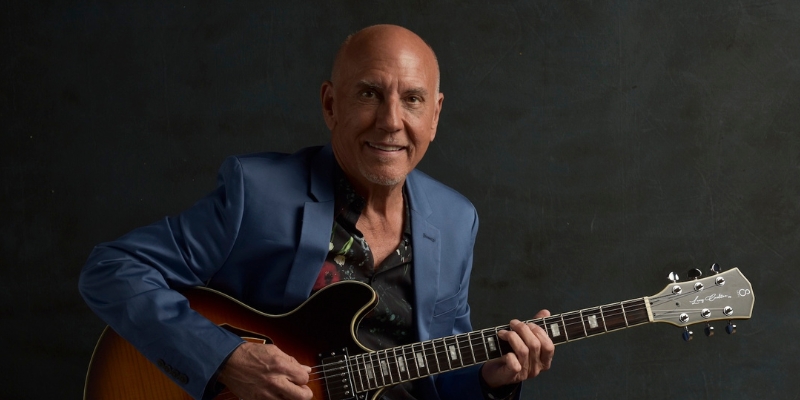 Larry Carlton (wearing a blue blazer) smiling and posing while holding a guitar