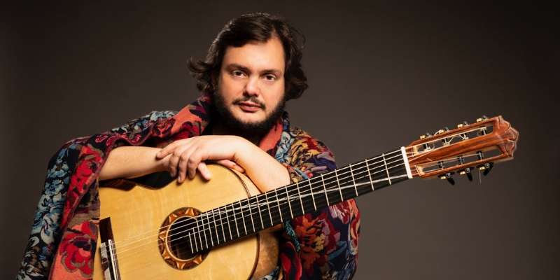 Yamandu Costa, wearing a colorful robe and posing with an acoustic guitar.